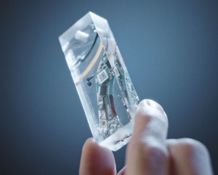 A hand holds the tech slice, a small cuboid of clear resin that contains parts relating to the Dyson OnTrac headphones.